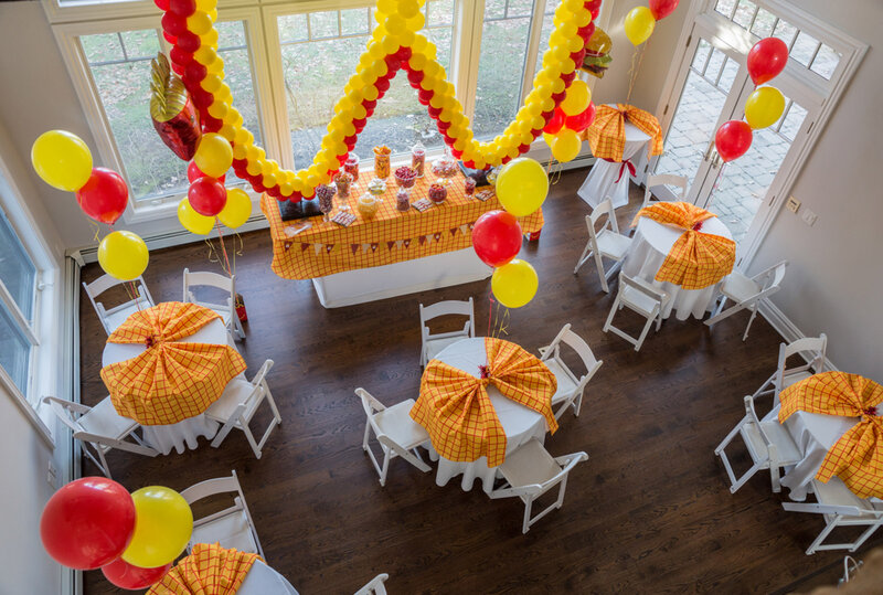 Room deocrated with yellow and orange balloons, with round tables set with orange and white table cloths